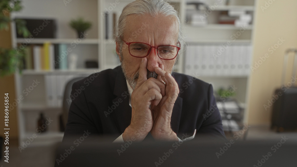 Fototapeta premium Man in suit with hand on chin and fingers touching nose, red glasses, seated at desk in building; thoughtful reflection.