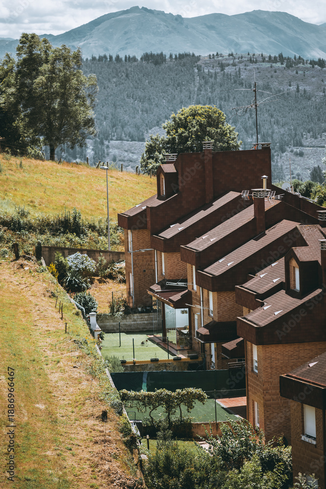 Fototapeta premium Telephoto view of a row of modern brick townhouses with brown tile roofs on a green hillside, fenced yards and small court, trees and dramatic mountain range in background