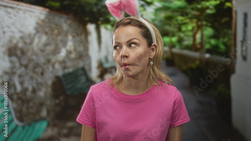 Φωτογραφία Woman with bunny ears headband and pink tshirt makes fish lips gesture on street beside a stone wall and bench; playfulness