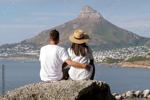 Bild auf Leinwand A couple sits on a rocky outcrop, embracing as they admire the breathtaking view of Table Mountain and the ocean