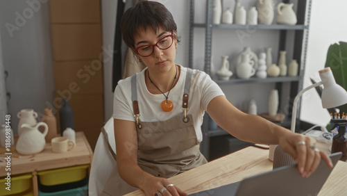 Fotografi Young woman in apron and glasses types on laptop with both hands beside finished ceramic vases and tools on table in pottery studio; concentration
