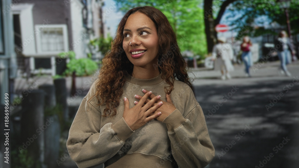 Fototapeta premium Young african american woman smiling with hands over chest on a city street wearing a beige sweatshirt and natural curls; gratitude warmth.