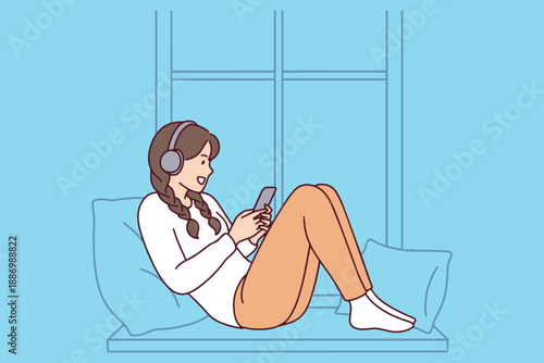 Mysterious young woman sitting by window using smartphone and wearing headphones. 