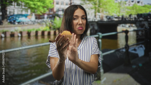 Photography Young hispanic woman holding a cupcake with hands pushing it away on a street in amsterdam; disgust rejection