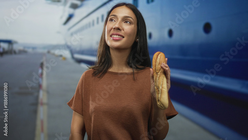 Cuadro en lienzo Young woman holding baguette and smiling with raised hand beside moored ship hull at building pier under daylight; travel joy