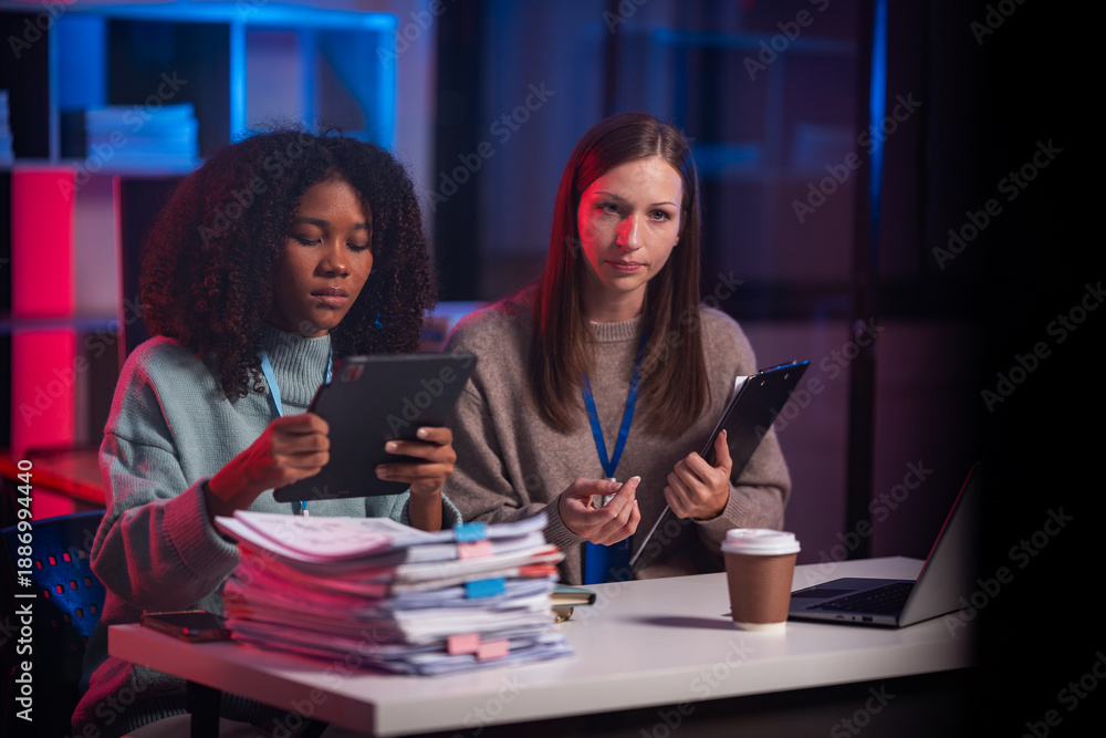 Fototapeta premium Two female employees work overtime late into the night handling documents and digital equipment in a modern office.