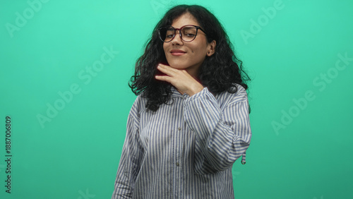 Valokuva Young arab woman with curly brunette hair and glasses touching cheek with hand in green studio wearing striped shirt and buttoned collar; calm confidence