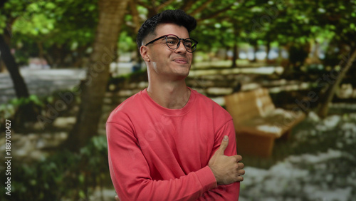 Young man smiling in a park wearing glasses and a red shirt with trees and benches in the background under natural daylight setting an outdoors scene