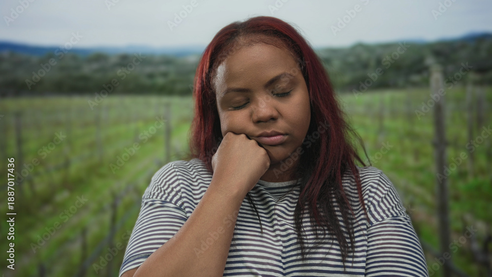 Obraz premium Woman resting head on hand with eyes closed in forest among vineyard stakes wearing striped shirt; exhaustion.