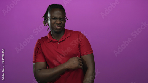Fotografie Young african american man grimacing while holding his arm in studio with purple backdrop; disgust reaction