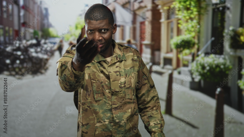 Obraz premium Man soldier in camouflage uniform beckons with hand toward camera, wearing backpack, closeup on a street; pride duty invitation.