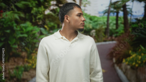 Man turns head to right on a shaded street path lined with palms and dense foliage, wearing cream polo and gold chain; calm reflection.