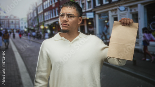 Young hispanic man holds paper bag aloft with right hand on a busy street, wearing glasses and cream sweater, inspecting the bag; urban confidence.