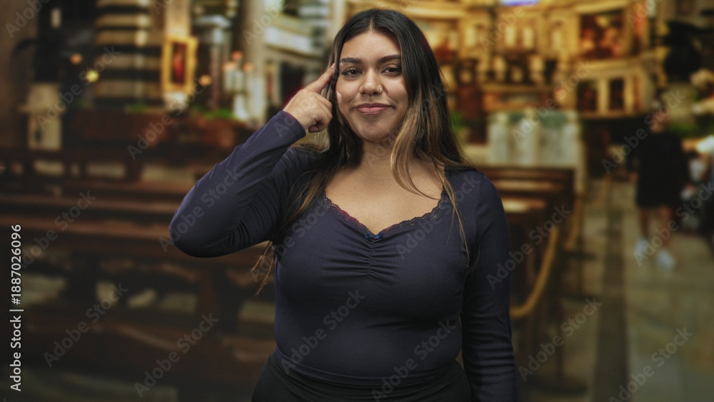 Fototapeta premium Woman pointing to temple while touching hair and smiling with closed eyes, wearing navy top and nose ring inside building; serenity confidence.