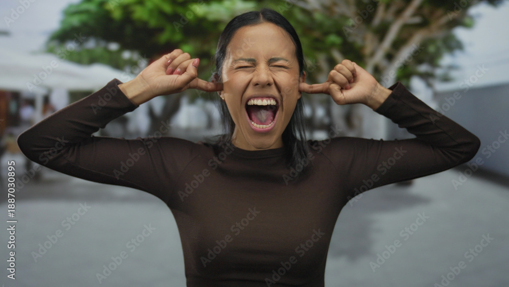 Naklejka premium Young hispanic woman outdoors screaming with eyes closed and fingers in ears, expressing frustration with blurred green trees in the background.