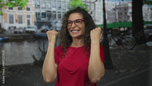 Foto Energetic hispanic woman raising clenched fists on urban street amid historic canal district with brick buildings in soft daylight; joy victory celebration empowerment