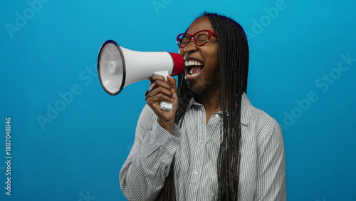 Woman shouting through megaphone against blue background wearing glasses and shirt with braids showing excitement and strength conveying communication and empowerment.