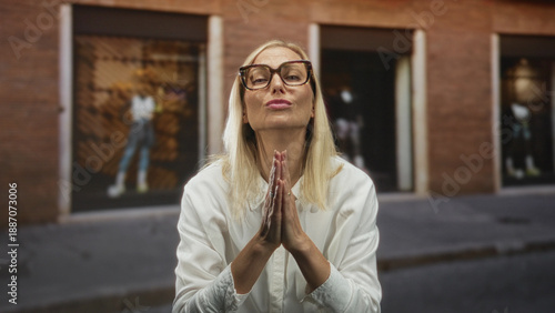 Fotografie Woman presses hands together in a prayer gesture, wearing white blouse and large glasses, standing on a city street in front of a shop building; hopeful plea