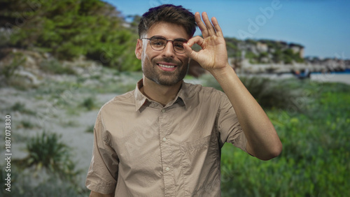 Man holds hand in ok sign near eye at beach under bright daylight by shore; serenity approval confidence.