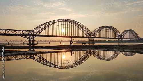 View of the sun creating a golden aura, reflecting on the water beneath the SaGaing bridge showcasing its intricate steel arches, SaGaing Bridge, Mandalay, Myanmar.