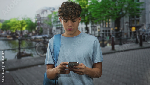 Tableau sur toile Teenage boy typing on smartphone, hands visible and looking down on street by canal and bicycles with backpack; quiet focus