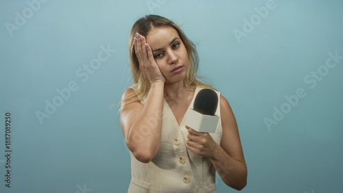 Slika na platnu Young blonde woman reporter holding microphone with hand on cheek in pale blue studio; tired resignation
