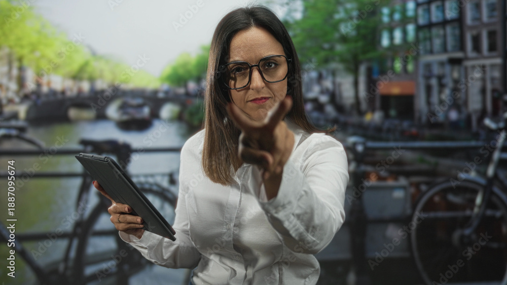 Fototapeta premium Woman holding tablet points finger to camera on a canal bridge street with bicycles and narrow buildings; determination challenge.