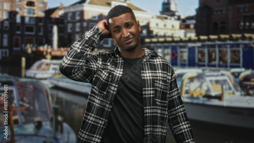Fotografie Young man scratches head, hand to temple gesture before canal boats backdrop in studio; uncertainty contemplation