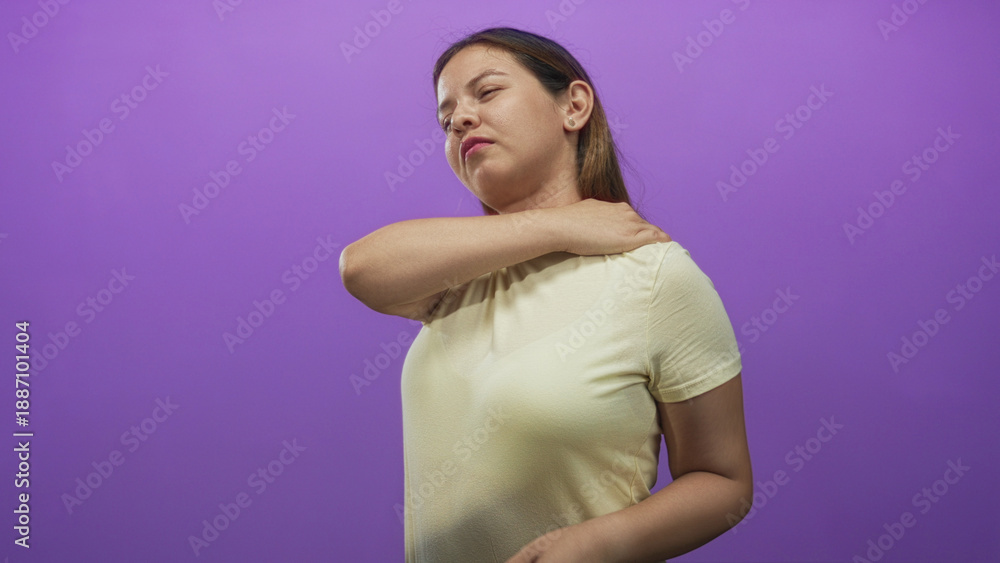 Obraz premium Woman rubbing her shoulder with hand, eyes closed, wearing yellow t shirt in purple studio; muscle tension discomfort.