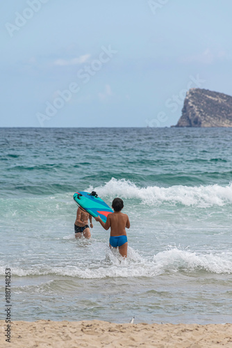 Two unrecognizable children play in the waves at a sandy beach, one holding a bodyboard, with gentle surf, turquoise water, and Benidorm Island visible on the horizon under a calm sky.