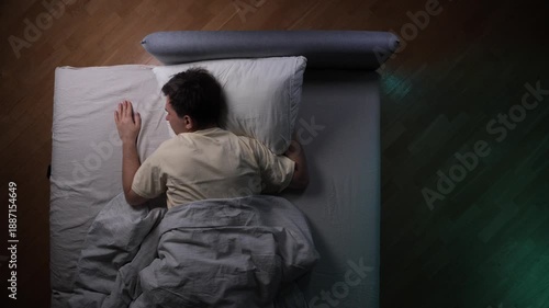 Reluctant man lying in bed, extending his arm to throw a ringing smartphone off the mattress, feeling annoyed and trying to avoid waking up in the morning