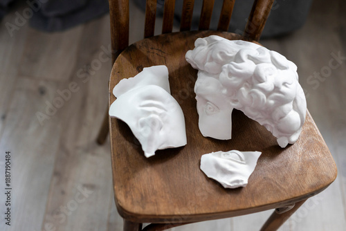 Fragmented pieces of a white plaster male head sculpture lying on an old wooden chair, creating an artistic and dramatic still life.