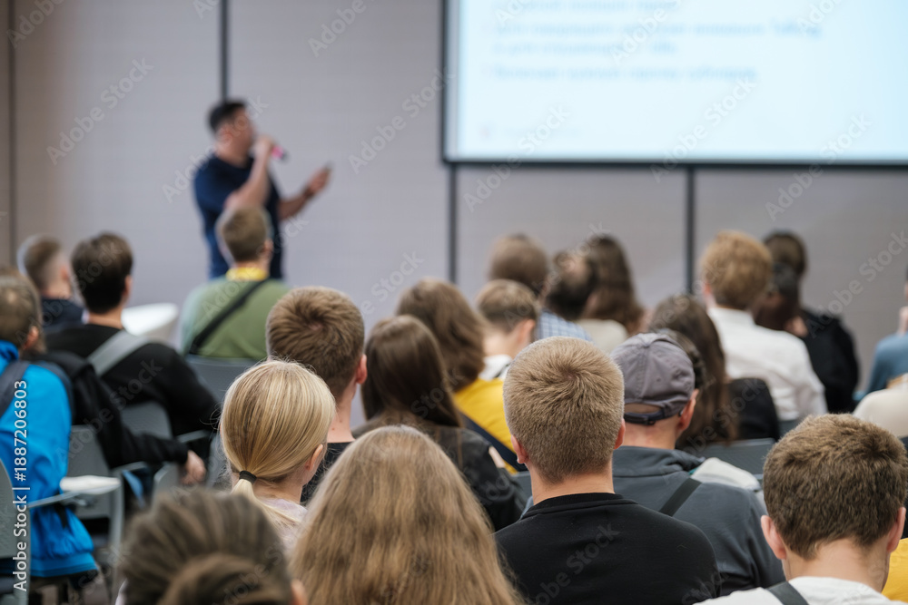Fototapeta premium Live presentation in front of attentive audience in conference setting. Speaker explains slides and engages crowd through talk visuals in classroom style venue.