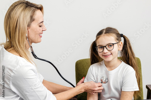 Tableau sur toile Female pediatrician using stethoscope to examine smiling girl with glasses sitti