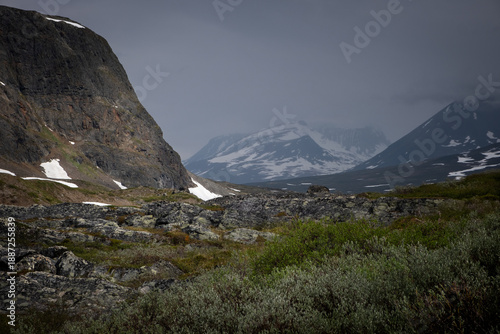 Dark, dramatic light falls on a steep rocky mountainside and tundra foreground along the Kungsleden trail in Swedish Lapland, while distant snow‑covered peaks fade into mist