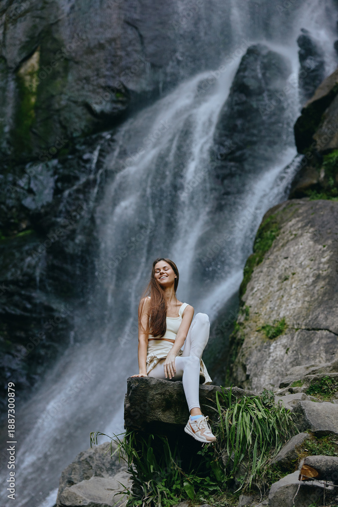 Obraz premium young woman sitting by waterfall in nature, wearing white clothes and sneakers, enjoying serene outdoor moment with greenery and rocks around, peaceful and relaxed atmosphere