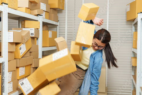 Asian adult woman small business owner losing balance while parcel boxes fall around warehouse aisle, ecommerce storage overload creating hazardous situation for online seller