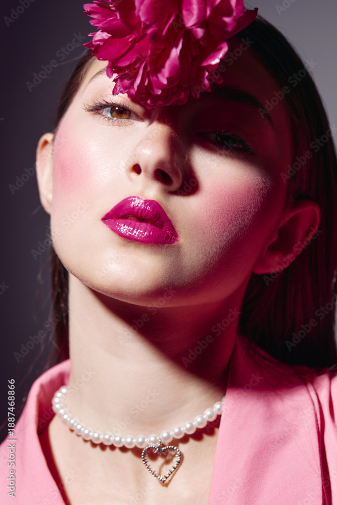 Fototapeta premium Portrait of a stylish woman with bright makeup and a large flower on her forehead. She wears a pearl necklace and pink outfit. Creative beauty and high fashion concept.
