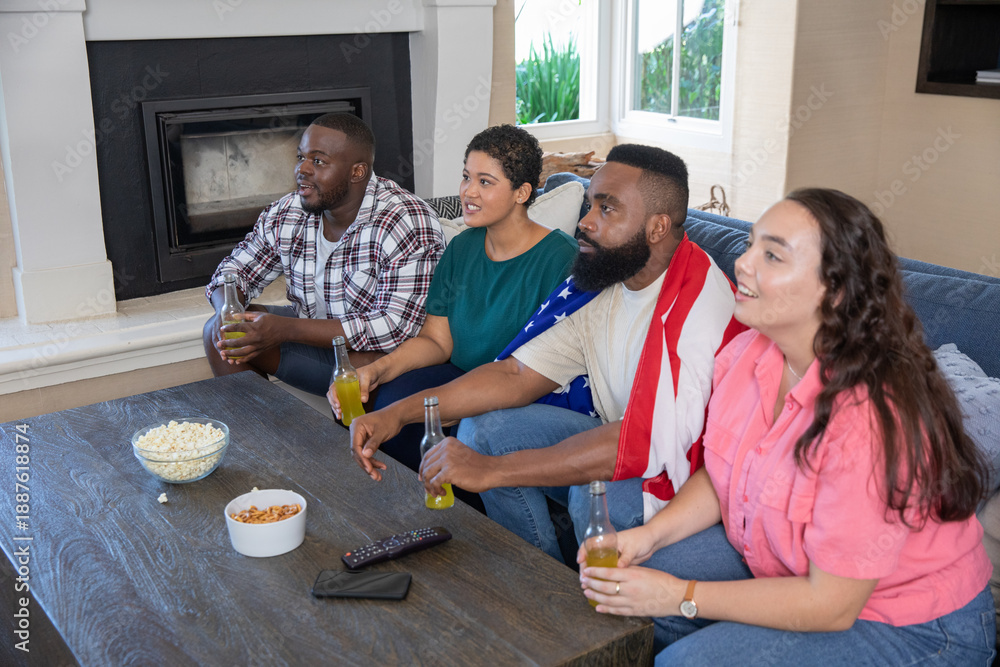 Obraz premium Diverse friends on sofa in living room and wearing american flag holding drinks, snack bowls