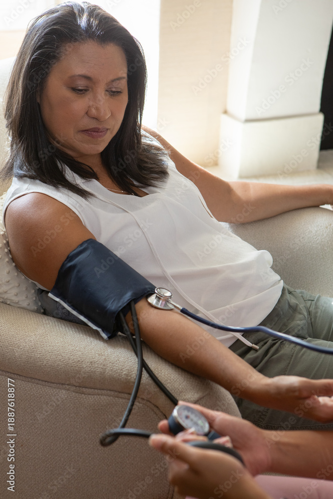 Naklejka premium Hispanic woman patient sitting in chair at home by window while nurse taking blood pressure gauge