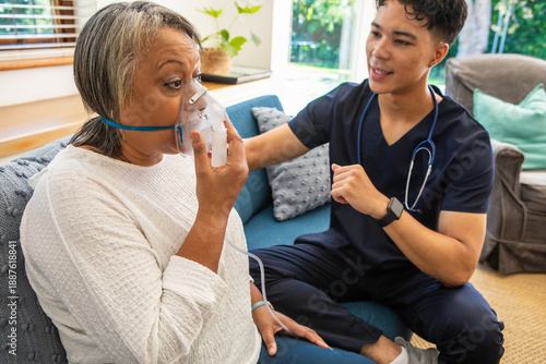 Senior african american patient and asian male nurse in scrubs sitting on sofa with oxygen mask