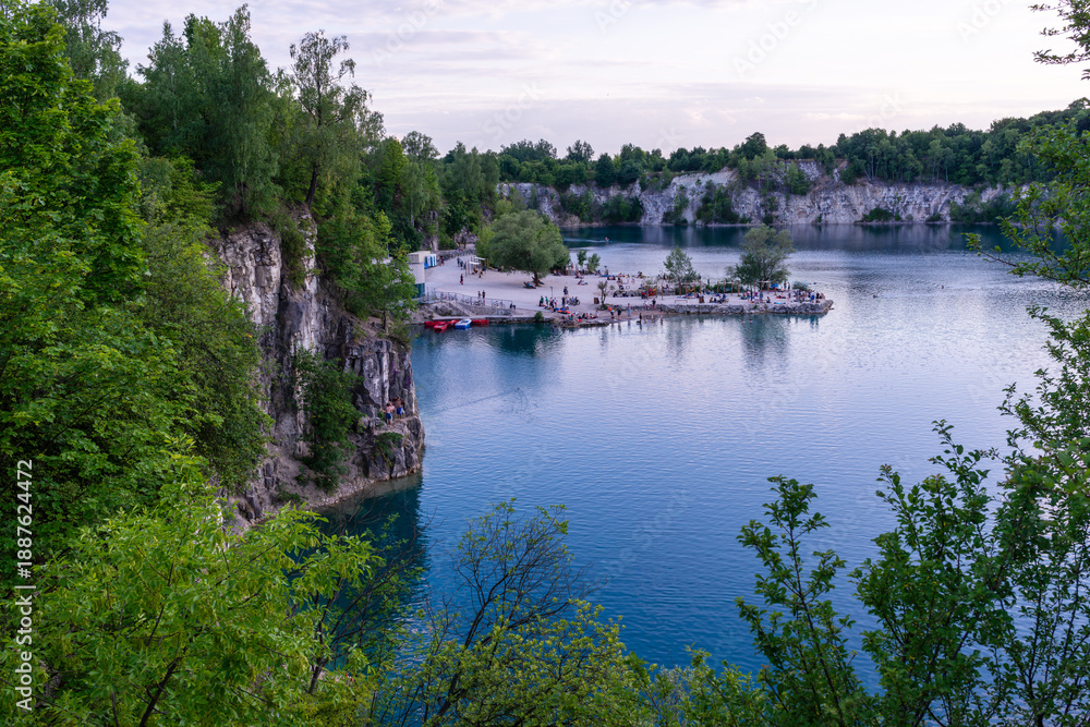 Fototapeta premium A panoramic view of Zakrzowek quarry lake near Krakow, showing steep cliffs, clear blue water, and a small recreational area with visitors along the shore. The scene captures summer leisure, nature to
