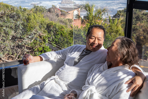 Diverse senior couple in bathrobes relaxing on couch at home holding glass tumblers