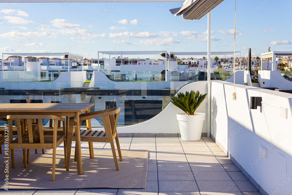 Naklejka premium Modern rooftop terrace with wooden dining set and glass railing in sunny residential complex