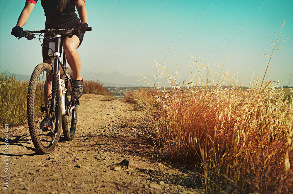 Naklejka premium Fitness, cycling and legs of person on bike for sports, training and challenge in nature. Exercise, workout and health with athlete on bicycle outdoor with gravel path, countryside or field for space