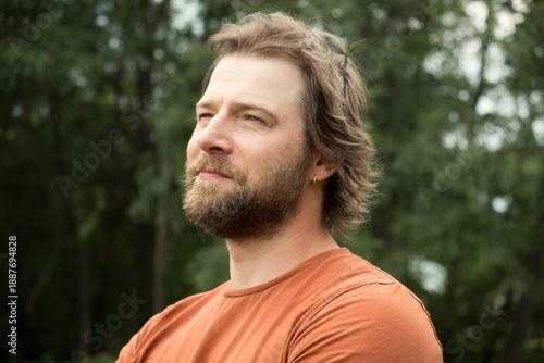 Portrait of handsome young man in orange t-shirt outdoors. Male with long hair and beard feel happy, calm and serious on Green nature background. Man looking away into distance. Diversity People