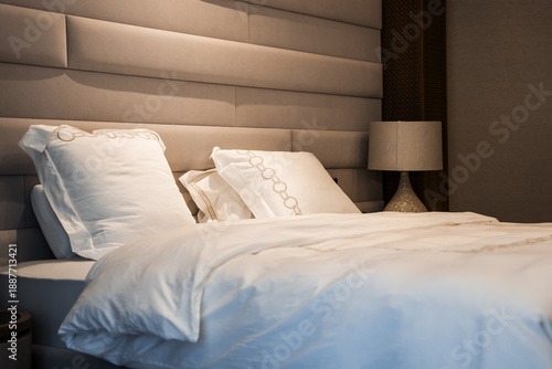 Neatly made bed with white linens rests under a paneled upholstered headboard. A ceramic bedside lamp sits by a woven accent panel, lit by warm evening ambient light.