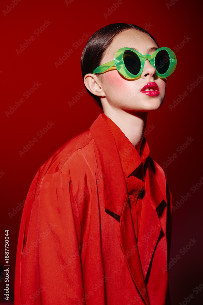 Fototapeta premium Fashionable young woman in oversized bright red coat and green round sunglasses poses confidently on a red background with bold makeup and vintage style. Creative beauty concept.