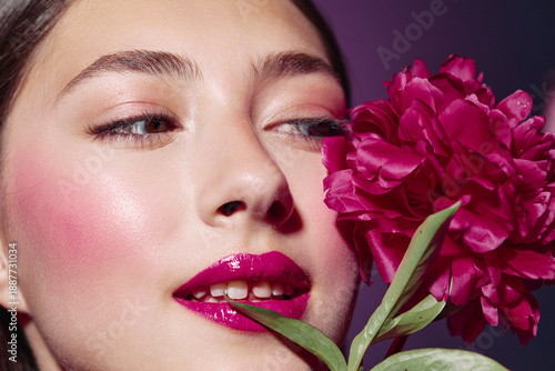 Close-up of a young woman with glossy fuchsia lipstick and blush posing with a vivid pink flower on purple background. Concept of bold beauty creative fashion style.