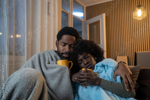 Young couple man and woman sitting on sofa at home wrapped in a blanket trying to warm up with hot cup of drink suffering from heating problems or feeling unhealthy with influenza.